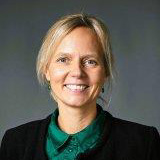 Portrait of a woman with blonde hair and black suit against a dark grey background.