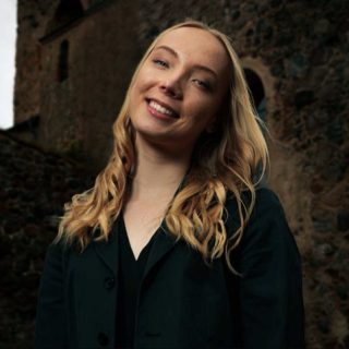 Person med ljust långt hår och svart blus mot mörk bakgrund.