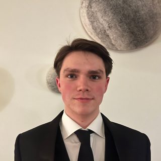 Person i svart kostym mot ljus bakgrund.