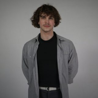 person med mörkt hår, iklädd mörk tröja och gr skjorta mot grå bakgrund.