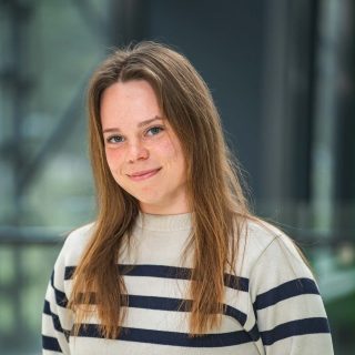 Person med långt hår och randig tröja mot grågrön bakgrund.