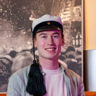 Person i studentmössa, vit t-shirt och grå tröja mot gråspräcklig bakgrund.