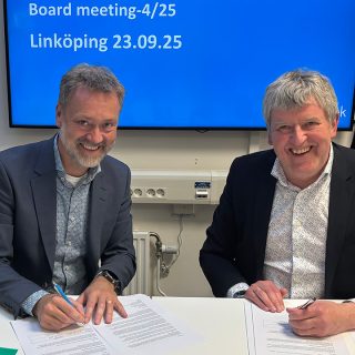 Two men sitting in front of a desk smiling and signing a paper
