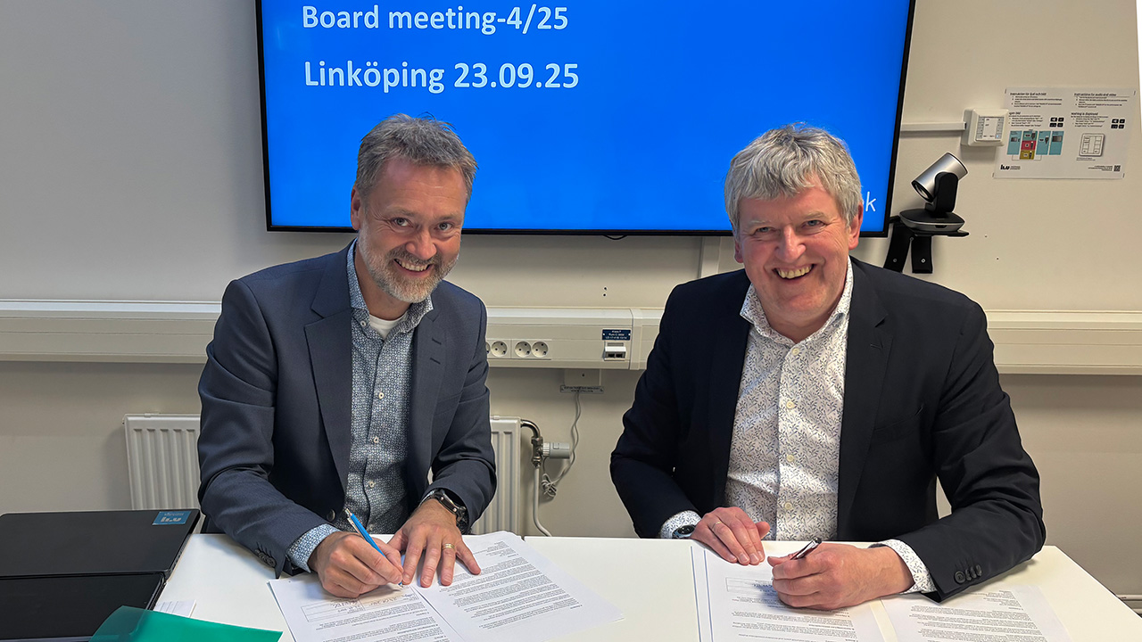 Two men sitting in front of a desk smiling and signing a paper