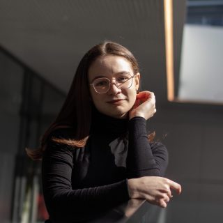 Person med långt mörkt hår, iklädd mörk långärmad tröja, lutar sig mot ett trappräcke.