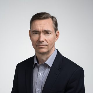 Man in black suit with light blue shirt infront of a light background.